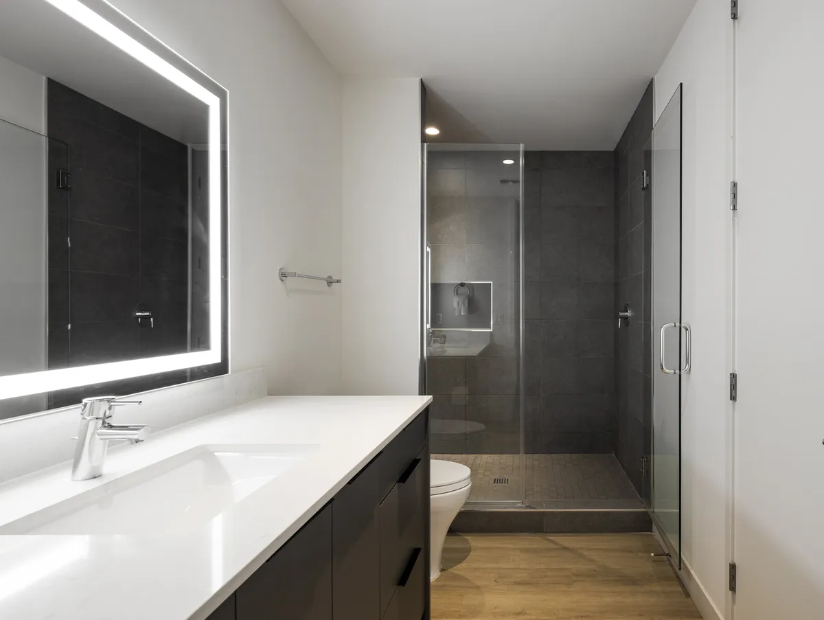 A modern, spa-style bathroom featuring a sleek vanity with an illuminated mirror, a glass-enclosed shower, and a toilet. The ambiance is clean, refreshing, and tranquil—ideal for self-care routines.