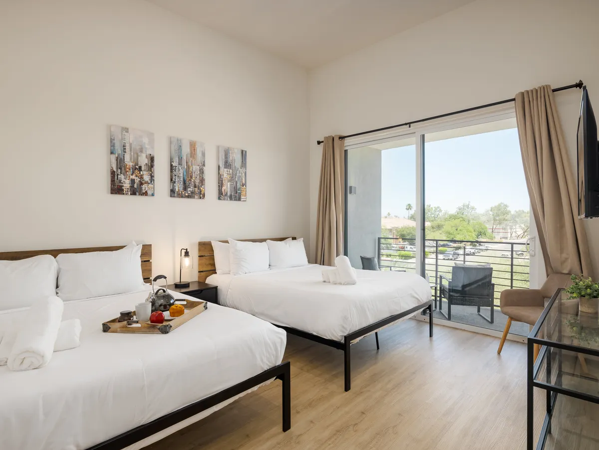 A bright, serene bedroom featuring two beds with white linens, decorative wall art, a sliding glass door leading to a private balcony, and a small dining table with a fruit tray. The natural light and balcony access add to the feeling of spaciousness and tranquility.