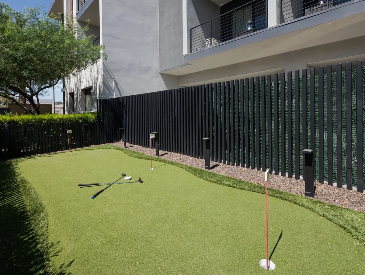 A modern putting green bordered by a green hedge and contemporary architecture. Golf clubs are visible, and the area invites guests of all ages for playful competition or casual relaxation.